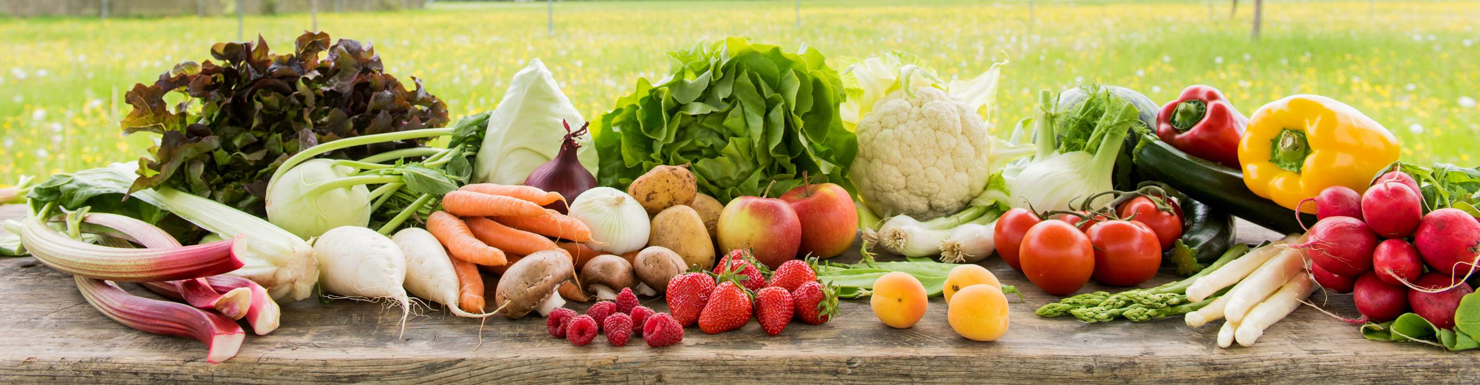 Frisches Obst und Gemüse, wie Salat, Rettich, Kartoffeln, Erdbeeren, Tomaten und Aprikosen auf einem Holztisch im Freien.