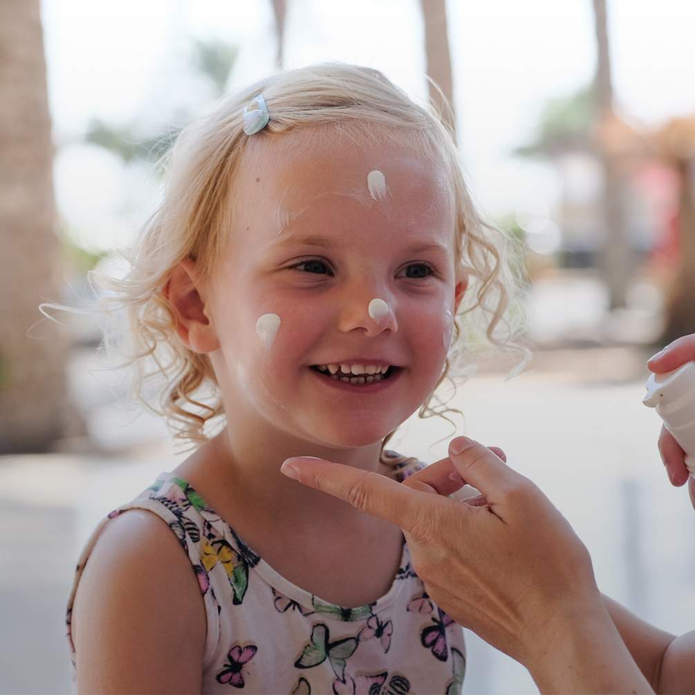 Kleines lächelndes Mädchen hat auf Nase, Wangen und Stirn weiße Sonnencreme-Punkte, die Hände der Mutter halten eine Packung Sonnencreme.