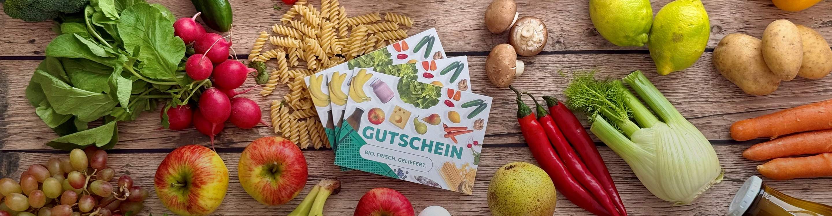Drei Isarland Gutscheinkarten inmitten von Produkten aus dem Isarland Sortiment, wie Obst, Gemüse, Brot, Käse, Eier und Nudeln.