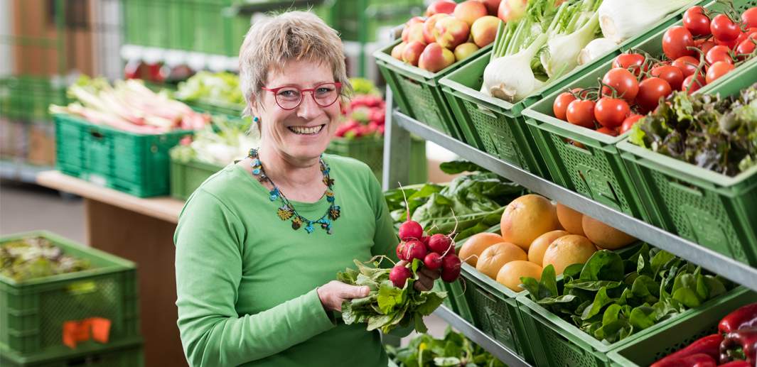 Lachende Ökokisten Mitarbeiterin mit Radieschenbund in der Hand steht vor einem Regal mit frischem Obst und Gemüse.