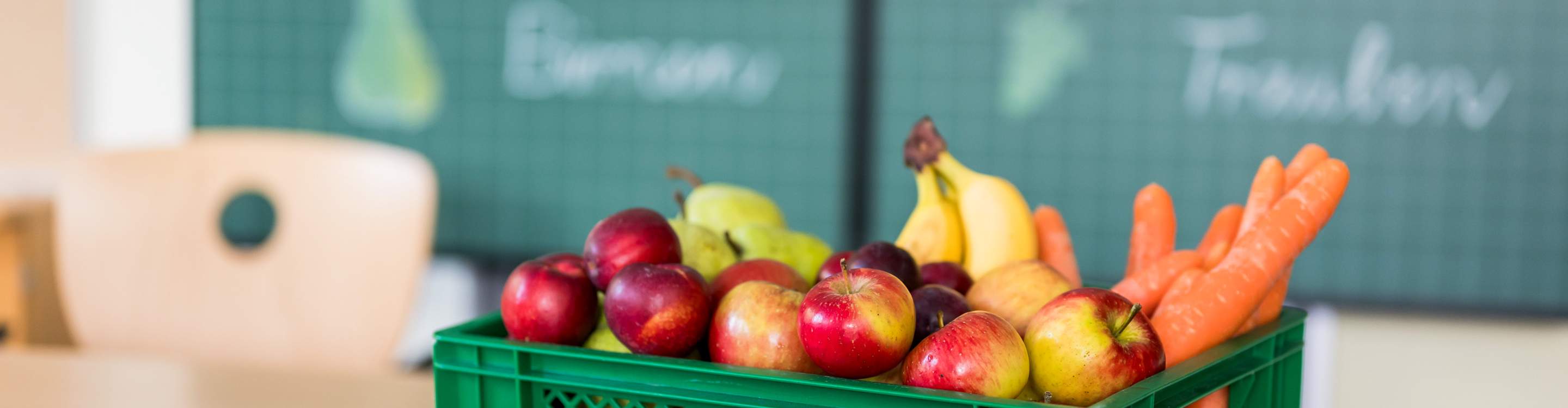 Eine Ökokiste, befüllt mit Äpfeln, Bananen, Birnen, Karotten und Nektarinen, steht auf dem Tisch in einem Klassenzimmer. Im Hintergund eine grüne Tafel.