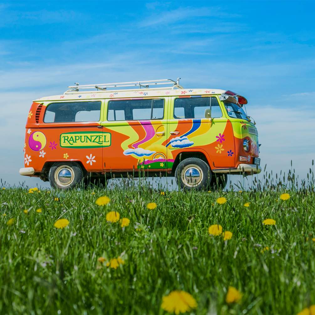 Ein Hippie-VW-Bus mit Rapunzel Logo steht auf der grünen Wiese.