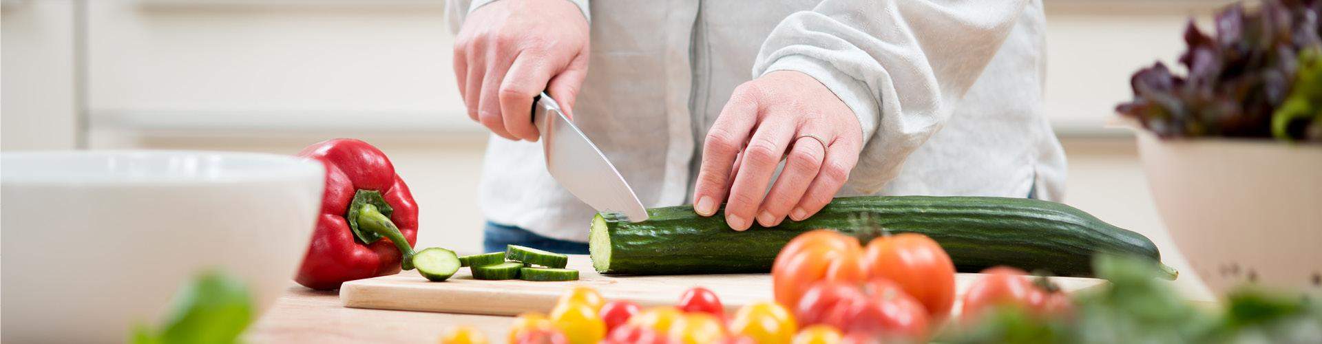 Frau schneidet Gurke auf einem Holzbrett, davor und daneben liegen Paprika und Tomaten.