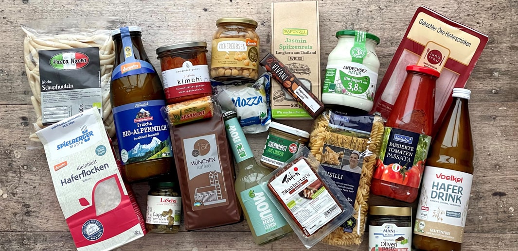 Viele Bio Produkte, wie Haferflocken, Mozzarella, Kaffee, Limo, Joghurt und Schinken auf einem Holztisch.