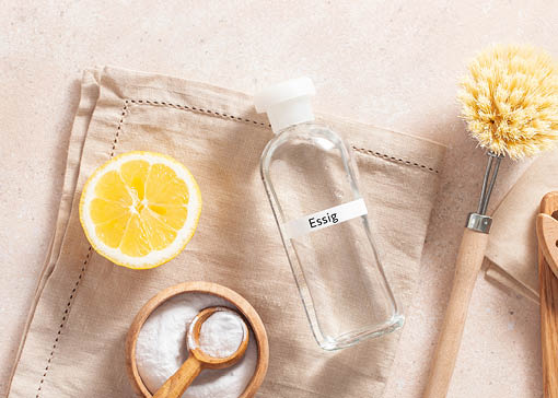 Arrangement auf beigem Geschirrtuch bestehend aus aufgeschnittener Zitrone, Holzschale mit Natron, leerer Flasche und Geschirrbürste.