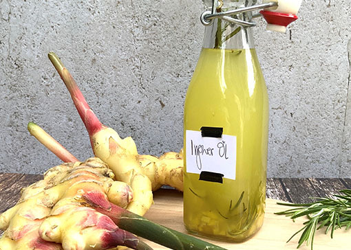 Arrangement aus kleiner Glasflasche mit gelblichem Ingweröl, frischen Ingwerknollen und Rosmarin auf einem Holztisch.