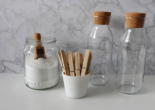 Ein offenes Glas mit Natron und Holzschäufelchen, ein Becher mit Holzwäscheklammern und zwei leere Glasflaschen mit Korkverschluss.