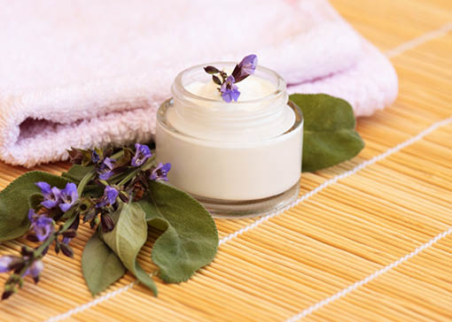 Arrangement auf Bambusmatte mit weißem Handtuch, Salbeiblüten und -blätter und Tiegel mit Creme.