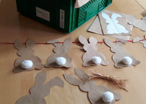 Aus Tütle-Papier ausgeschnittenen Hasen mit weißem Puschel an roter Schnur liegen auf einem Tisch. Im Hintergrund ist die grüne Ökokiste zu sehen.