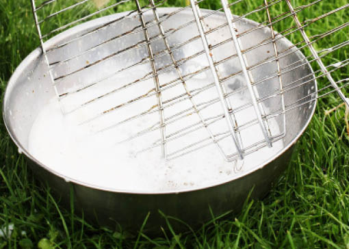 Metallschüssel gefüllt mit schaumigem Wasser steht auf einer Wiese, in der Schüssel liegt ein Fischgrillrost.