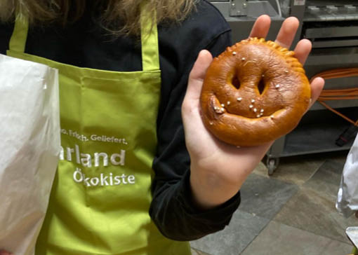 Kind mit grüner Isarland Schürze hält selbstgebackene Smiley-Brezensemmel in die Kamera.