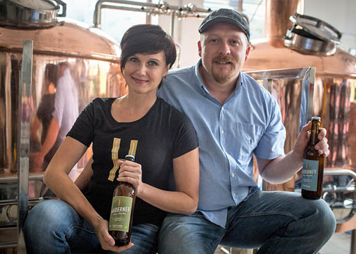 Thomas und Marta Girg sitzen vor einem Kupferkessel mit jeweils eine Flasche Bier in der Hand.