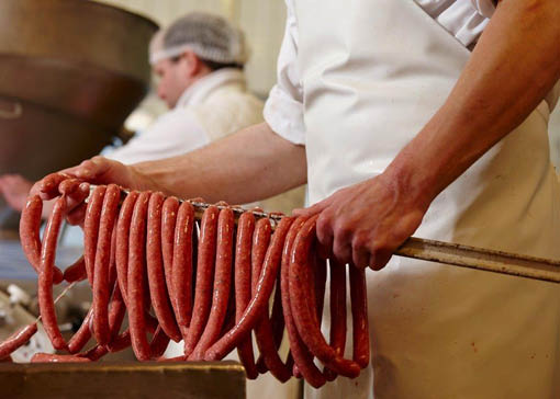 Ein Metzger hält einen Holzstab in der Hand. Auf dem Stab hängen Wurstpaare.