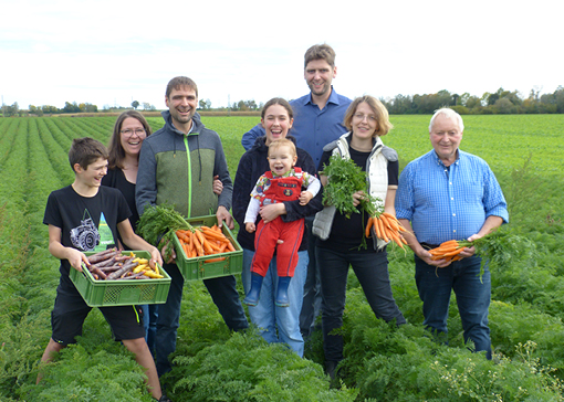 Familie Pfänder steht auf einem Feld. Manche von ihnen halten einen Karottenbund oder eine Kiste mit verschiedenfarbigen Karotten in den Händen.