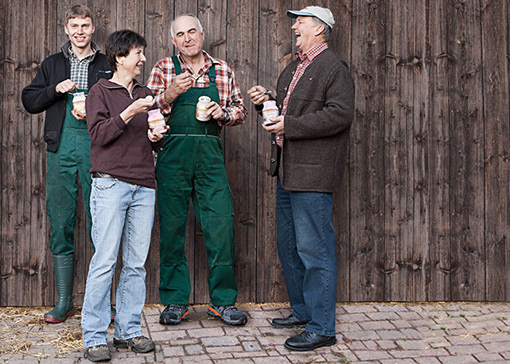 Eine Bäuerin und drei Bauer halten Joghurtgläser in der Hand, essen und lachen.