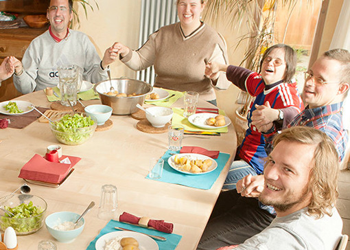 Einige Personen sitzen um einen Tisch, halten sich Hände und lachen.