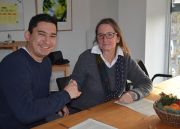 Sternekoch Tohru Nakamura und Geschäftsführerin der Isarland Ökokiste sitzen handeschüttelnd und lächelnd an einem Tisch.