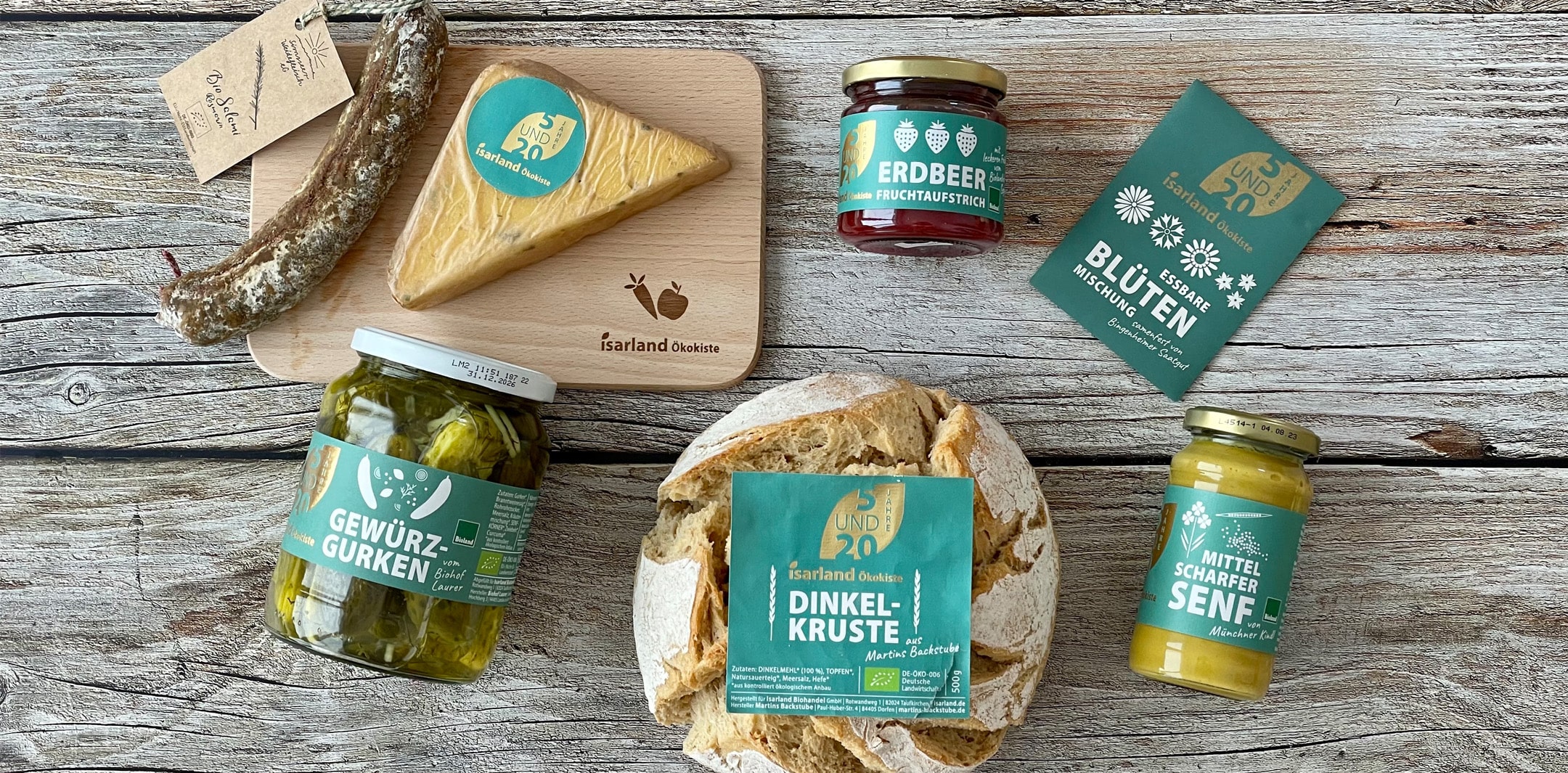 Mit dem Jubiläumslogo gelabelte Isarlandprodukte, wie Salami, Käse, Gewürzgurken, Marmelade, Brot, Blütensamen und Senf auf einem Holztisch.