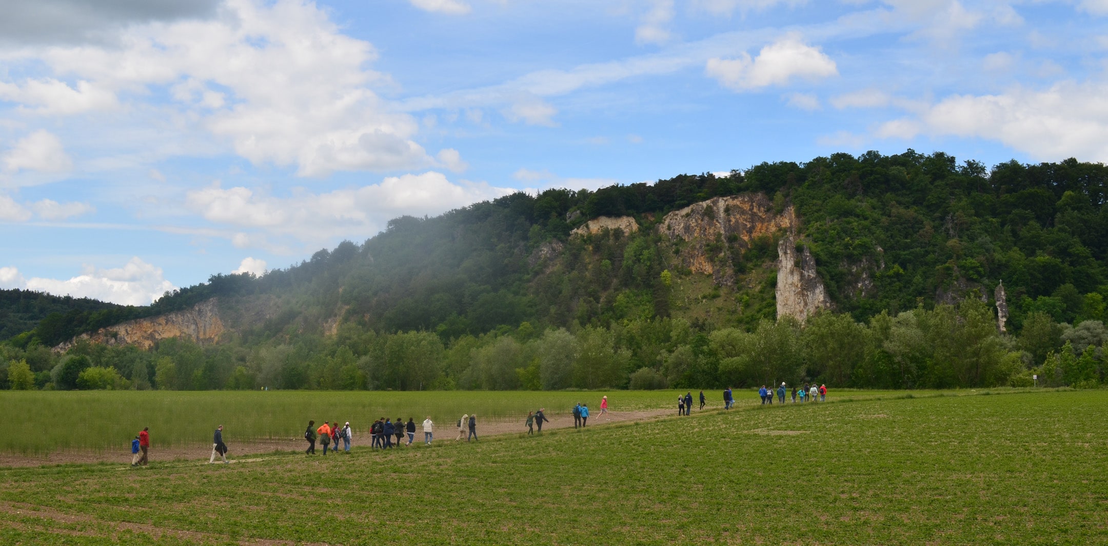 Besuchende der Führung am Biolandhof Keil spazieren in der Ferne auf einem Feldweg, vor bewaldeten Kalksteinhängen.