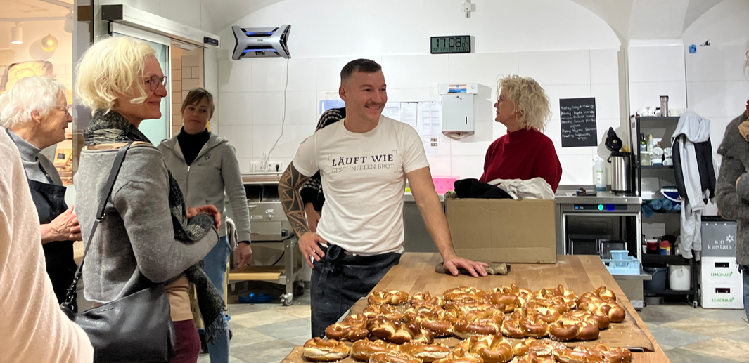 Bäckermeister Nico Scheller steht lächelnd mit Besucherinnen und Besuchern in seiner Backstube vor einem Tisch mit frisch gebackenen Brezen.